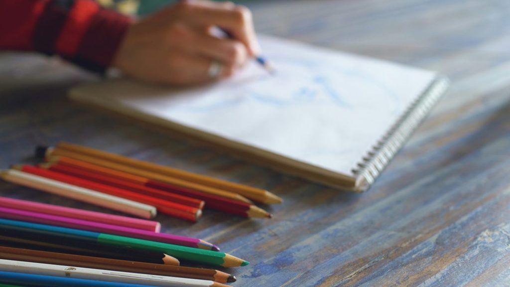 Artiste dessinant dans un carnet avec des crayons de couleur à proximité.